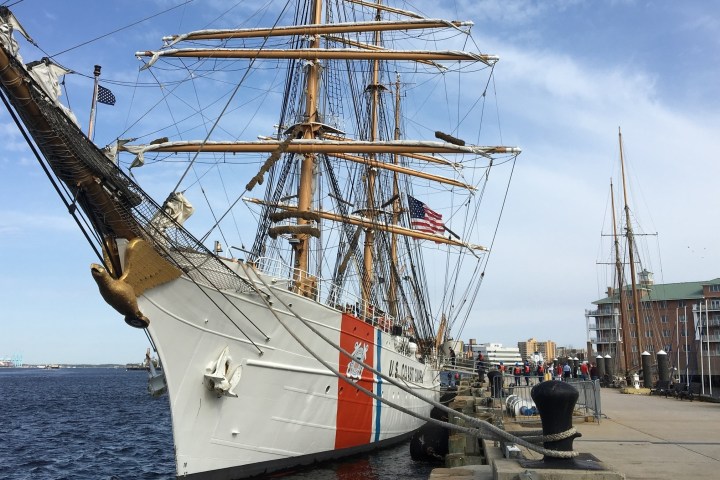 Historic tall ship docked at harbor with people nearby and buildings in background.