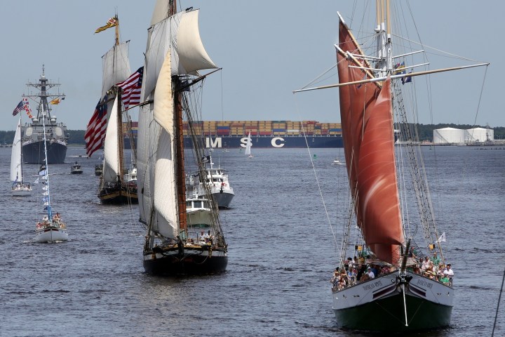 Sailboats and ships on a river with a container ship in the background.
