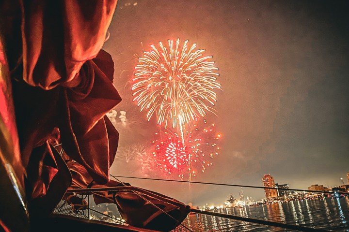 Fireworks over water at night, view from a boat with city skyline in background.